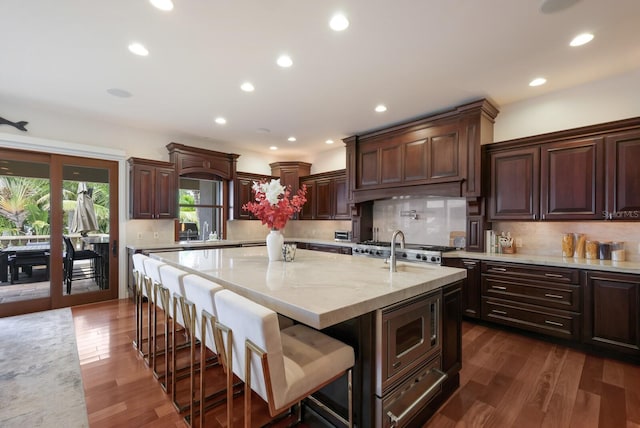 kitchen featuring stainless steel microwave, an island with sink, backsplash, and dark wood-type flooring