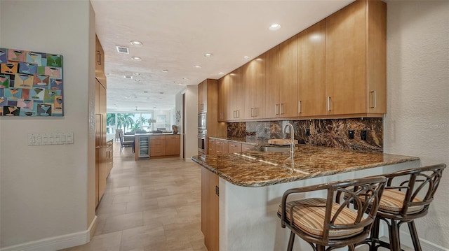 kitchen featuring a kitchen bar, sink, kitchen peninsula, dark stone counters, and beverage cooler