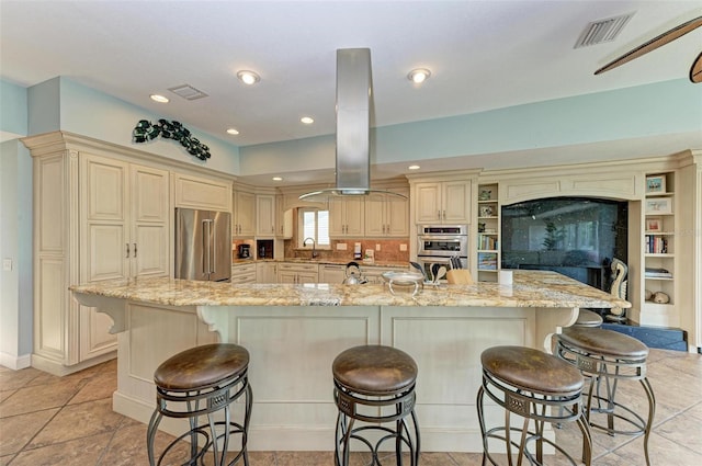 kitchen with cream cabinetry, appliances with stainless steel finishes, a large island with sink, fume extractor, and light tile floors