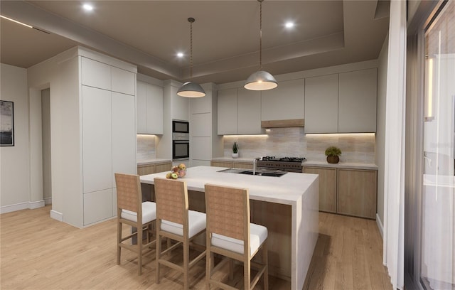 kitchen featuring tasteful backsplash, decorative light fixtures, a tray ceiling, a kitchen island with sink, and light wood-type flooring