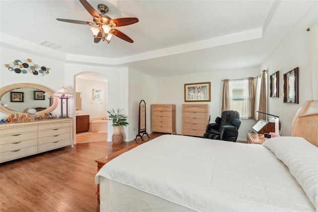 bedroom featuring a tray ceiling, connected bathroom, wood-type flooring, and ceiling fan