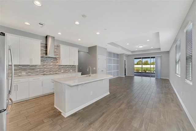 kitchen featuring white cabinetry, stainless steel refrigerator, a tray ceiling, an island with sink, and wall chimney range hood