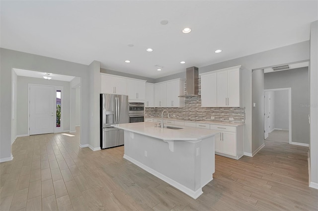 kitchen featuring wall chimney range hood, sink, appliances with stainless steel finishes, white cabinets, and a center island with sink