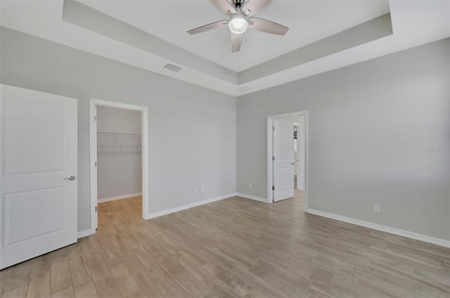 unfurnished bedroom with ceiling fan, a raised ceiling, a spacious closet, a closet, and light wood-type flooring