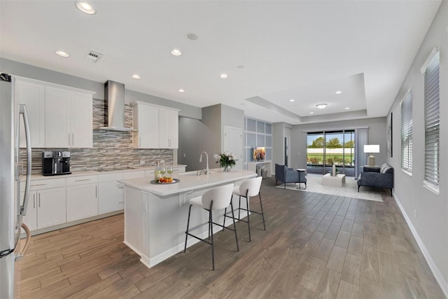 kitchen featuring wall chimney range hood, stainless steel fridge, a breakfast bar, a kitchen island with sink, and white cabinets