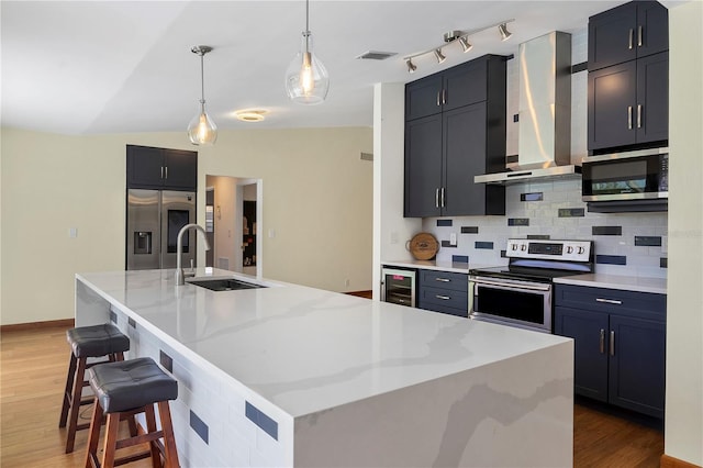 kitchen featuring a center island with sink, wall chimney range hood, appliances with stainless steel finishes, decorative light fixtures, and beverage cooler