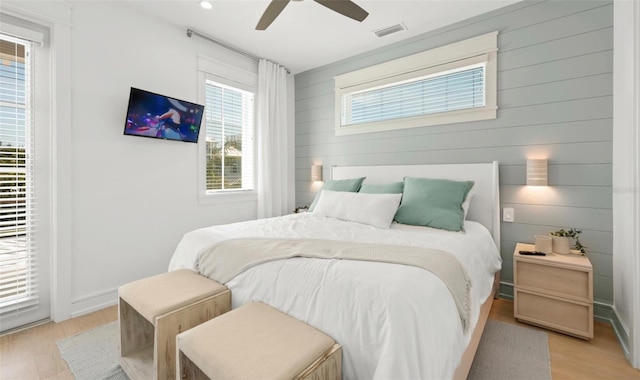 bedroom featuring multiple windows, light hardwood / wood-style flooring, ceiling fan, and wooden walls