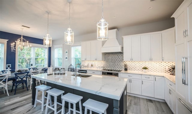 kitchen with decorative light fixtures, white cabinetry, premium range hood, and an island with sink