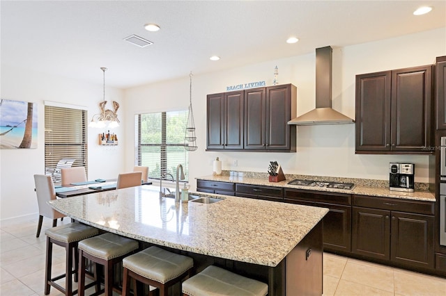 kitchen with stainless steel gas stovetop, sink, wall chimney exhaust hood, an island with sink, and dark brown cabinetry