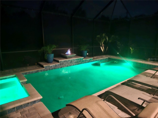 pool at night featuring glass enclosure, an outdoor fire pit, and a patio area