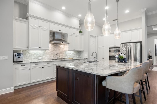 kitchen with white cabinetry, wall chimney exhaust hood, decorative light fixtures, a kitchen island with sink, and appliances with stainless steel finishes