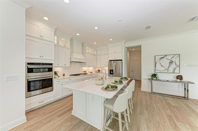 kitchen featuring premium range hood, white cabinetry, light hardwood / wood-style flooring, appliances with stainless steel finishes, and an island with sink