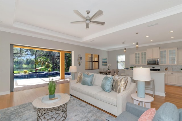 living room with ceiling fan, a raised ceiling, light wood-type flooring, and crown molding