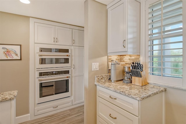 kitchen featuring white cabinetry, light stone counters, light hardwood / wood-style flooring, stainless steel double oven, and backsplash