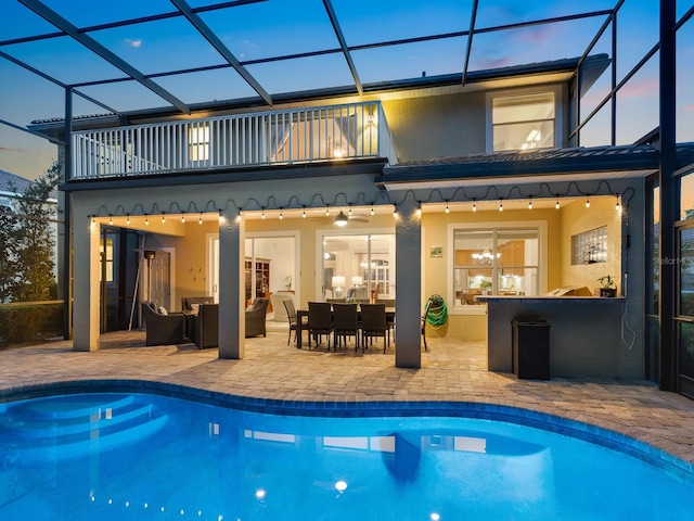 back house at dusk with a patio area, ceiling fan, a balcony, and glass enclosure