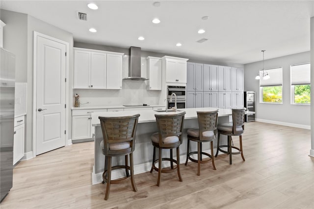 kitchen with light wood-type flooring, wall chimney range hood, pendant lighting, a center island with sink, and white cabinets