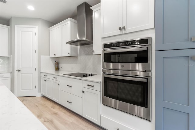 kitchen with stainless steel double oven, wall chimney range hood, black electric cooktop, white cabinets, and light wood-type flooring