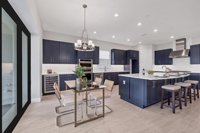 kitchen featuring a kitchen island with sink, wall chimney range hood, pendant lighting, a chandelier, and wine cooler