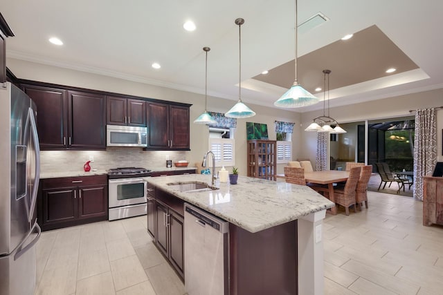 kitchen featuring stainless steel appliances, a raised ceiling, sink, decorative light fixtures, and an island with sink