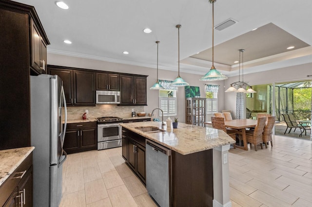 kitchen with a center island with sink, sink, appliances with stainless steel finishes, a tray ceiling, and decorative light fixtures