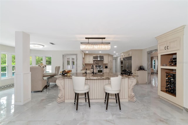 kitchen featuring a center island with sink, wall chimney range hood, hanging light fixtures, appliances with stainless steel finishes, and tasteful backsplash