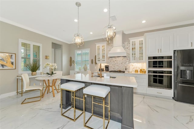 kitchen with premium range hood, stainless steel appliances, a kitchen island with sink, white cabinets, and hanging light fixtures