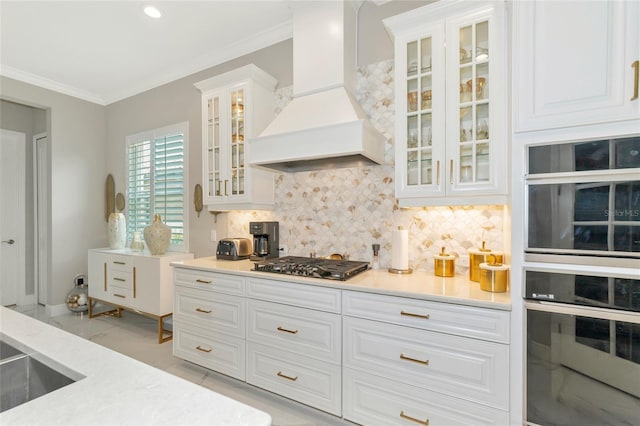 kitchen featuring backsplash, premium range hood, double wall oven, stainless steel gas cooktop, and white cabinetry