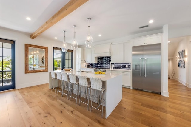 kitchen featuring custom exhaust hood, appliances with stainless steel finishes, beam ceiling, white cabinets, and a center island with sink