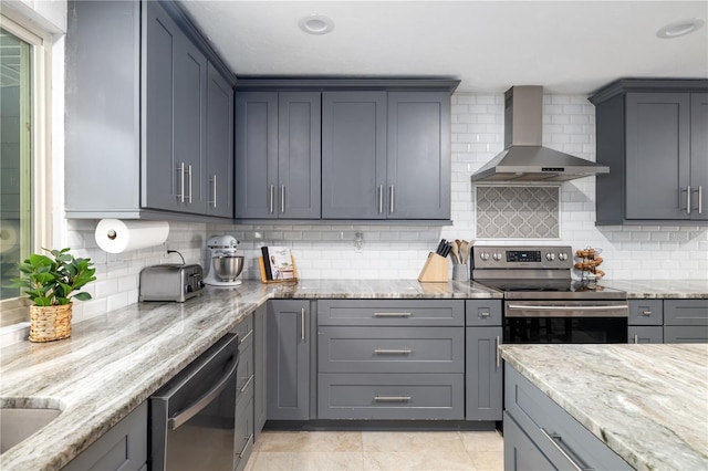 kitchen with stainless steel appliances, wall chimney range hood, gray cabinets, light stone countertops, and tasteful backsplash