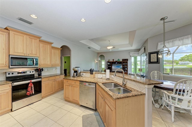 kitchen with sink, stainless steel appliances, an island with sink, a tray ceiling, and light brown cabinetry