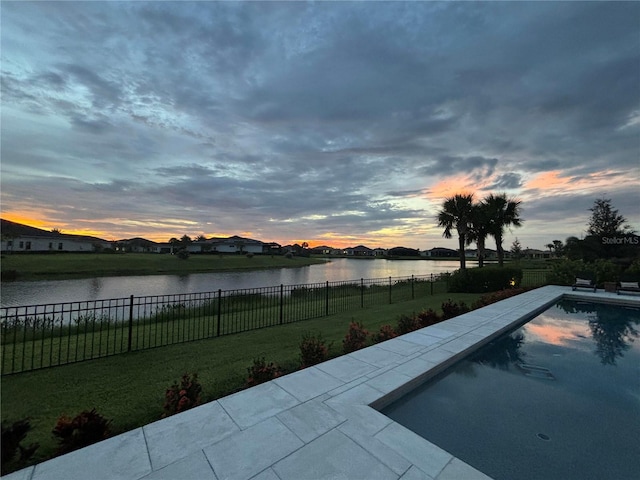 pool at dusk featuring a lawn, a water view, and a patio