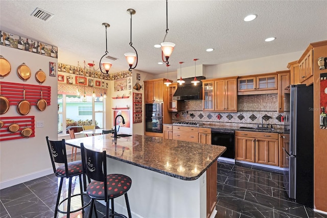 kitchen with decorative light fixtures, black appliances, dark stone countertops, a kitchen island with sink, and backsplash