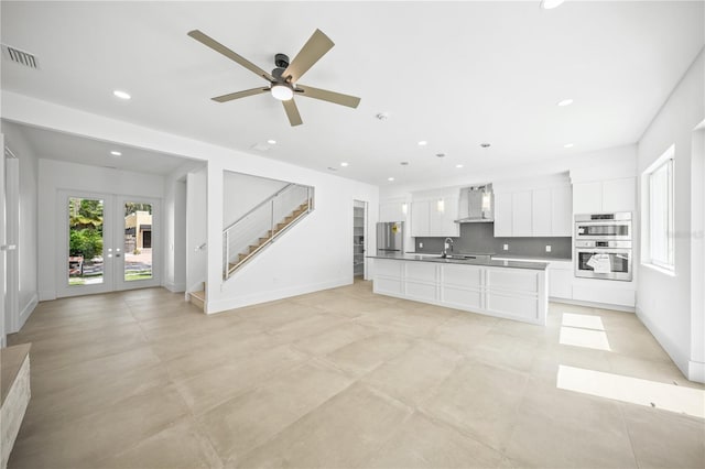 kitchen featuring a center island with sink, wall chimney range hood, hanging light fixtures, appliances with stainless steel finishes, and white cabinetry