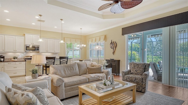 living room with ceiling fan with notable chandelier, a tray ceiling, a wealth of natural light, and light hardwood / wood-style floors