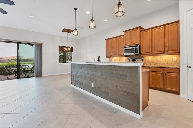 kitchen featuring decorative light fixtures, backsplash, a kitchen island with sink, light tile patterned floors, and light stone counters