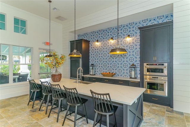 kitchen with an island with sink, sink, double oven, and decorative light fixtures