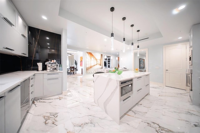 kitchen with a raised ceiling, hanging light fixtures, light stone countertops, a kitchen island, and white cabinetry