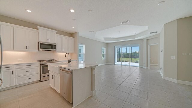 kitchen featuring appliances with stainless steel finishes, a center island with sink, light tile patterned floors, white cabinetry, and a raised ceiling