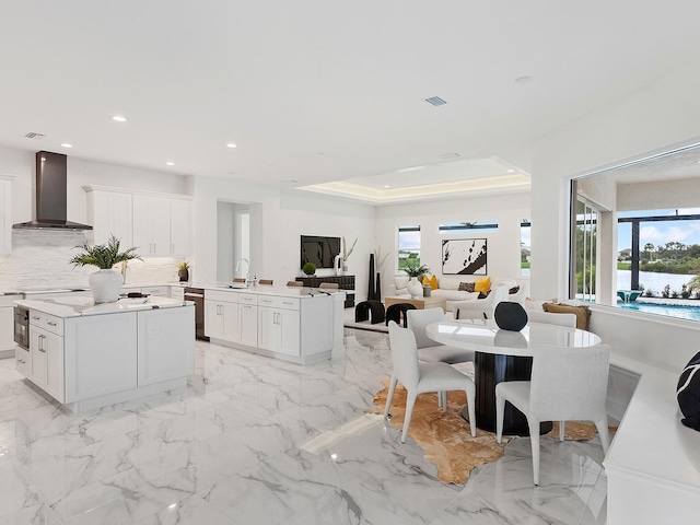 kitchen featuring a raised ceiling, white cabinetry, a kitchen island, and wall chimney range hood