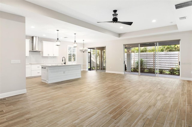 kitchen featuring ceiling fan with notable chandelier, wall chimney range hood, decorative light fixtures, white cabinetry, and an island with sink