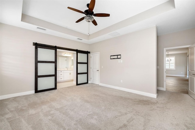 unfurnished bedroom with ensuite bath, a barn door, a tray ceiling, light carpet, and ceiling fan with notable chandelier