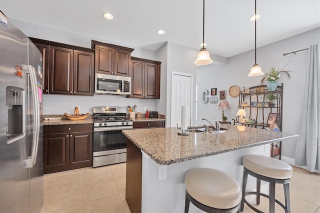 kitchen featuring appliances with stainless steel finishes, light stone counters, a kitchen island with sink, light tile patterned floors, and hanging light fixtures