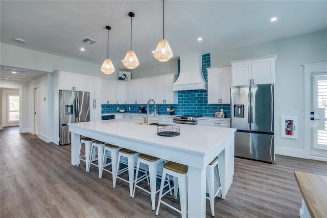 kitchen with hanging light fixtures, appliances with stainless steel finishes, an island with sink, white cabinets, and custom exhaust hood