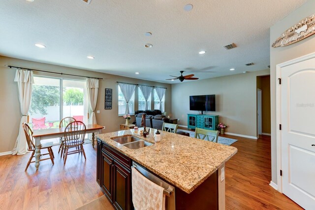 kitchen with ceiling fan, dishwasher, light stone countertops, a center island with sink, and light hardwood / wood-style flooring