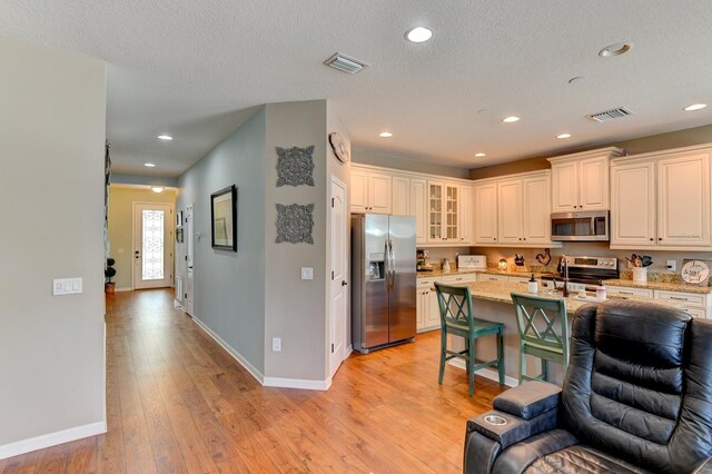 kitchen with light wood-type flooring, light stone countertops, appliances with stainless steel finishes, a textured ceiling, and a kitchen island with sink