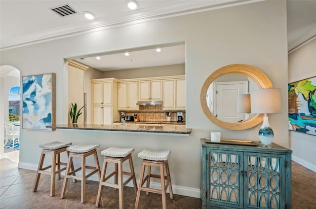 kitchen featuring cream cabinetry, kitchen peninsula, decorative backsplash, dark tile patterned flooring, and stone counters