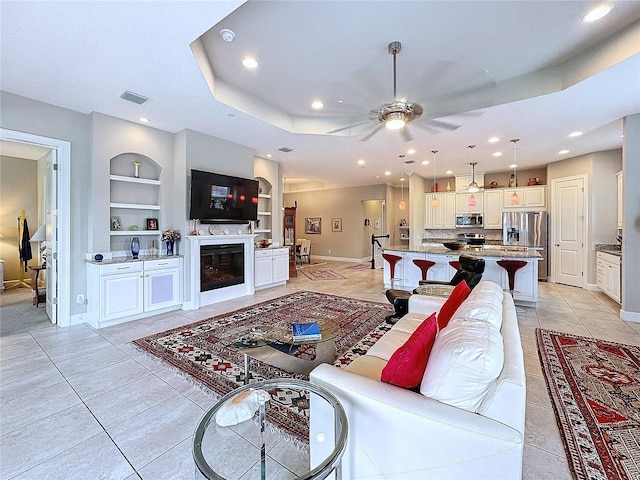 living room featuring built in shelves, light tile patterned floors, a raised ceiling, a glass covered fireplace, and baseboards