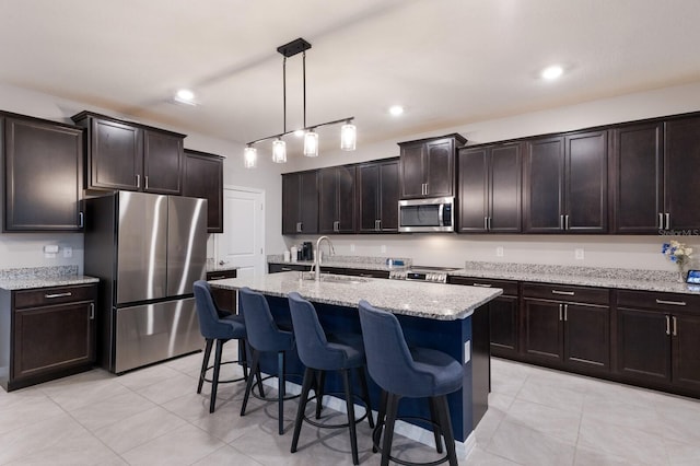 kitchen with decorative light fixtures, an island with sink, dark brown cabinetry, stainless steel appliances, and light stone countertops