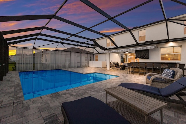 pool at dusk with a bar, outdoor lounge area, a patio, and glass enclosure