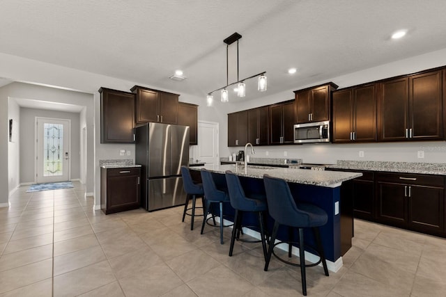 kitchen featuring appliances with stainless steel finishes, a kitchen island with sink, dark brown cabinetry, light stone countertops, and decorative light fixtures